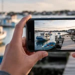 Hands hold an iPhone photographing boats at a harbor dock.